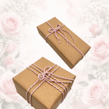 Two brown kraft paper gift boxes wrapped with pink and white twisted twine and tied with bows, set against a soft white background decorated with light pink roses.