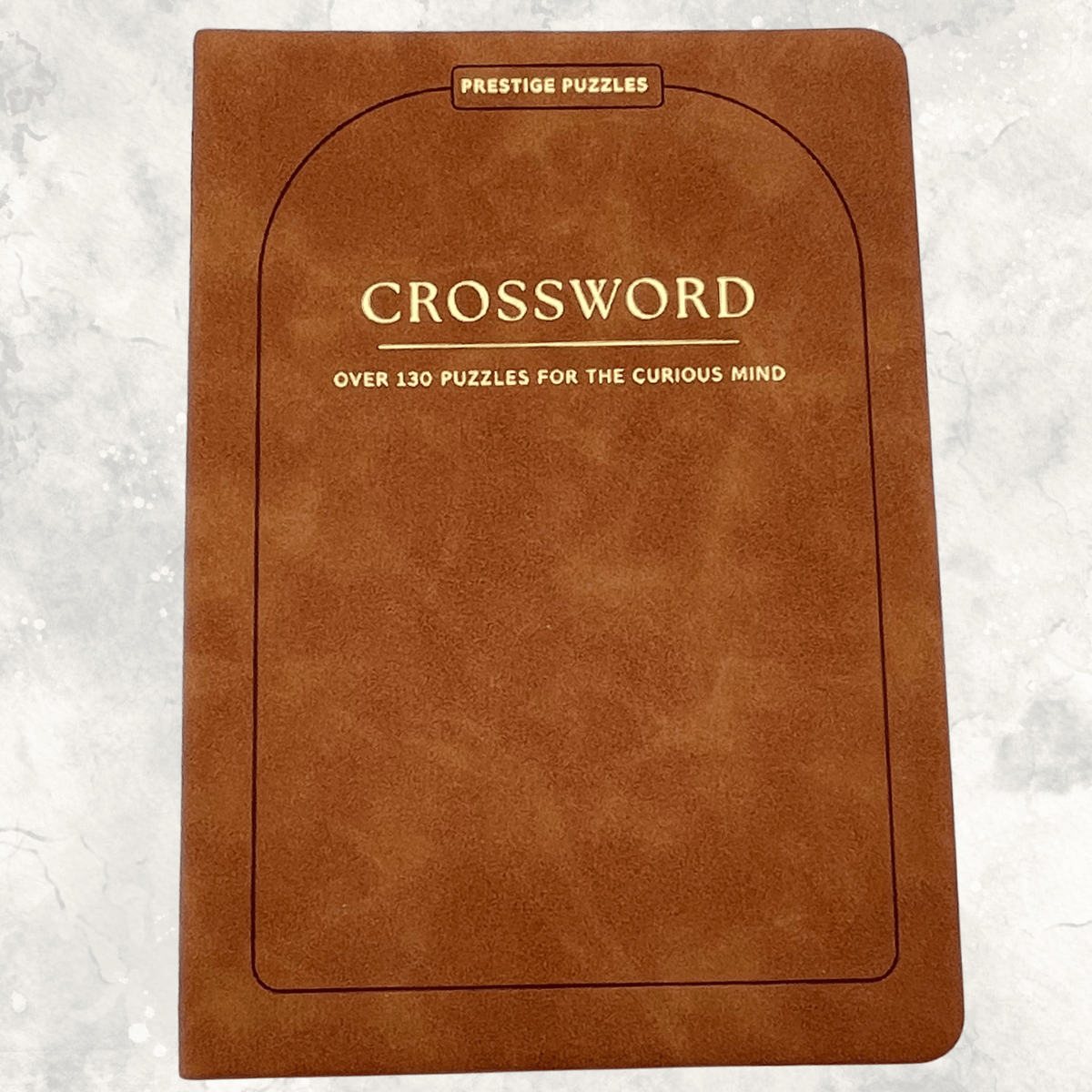 Tan-brown faux suede crossword puzzle book with gold lettering featuring over 130 crossword puzzles