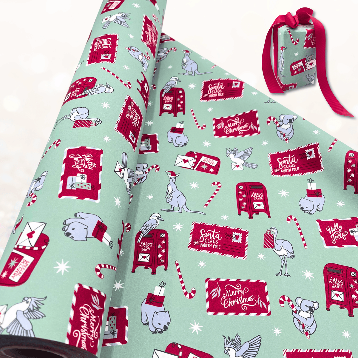 A roll of Christmas-themed wrapping paper is partially unrolled, showing a festive pattern of red post boxes, Santa mailers, candy canes, and koalas on a mint green background. A small wrapped gift box with a red ribbon sits to the upper right on a light, softly blurred surface.