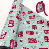 A roll of Christmas-themed wrapping paper is partially unrolled, showing a festive pattern of red post boxes, Santa mailers, candy canes, and koalas on a mint green background. A small wrapped gift box with a red ribbon sits to the upper right on a light, softly blurred surface.