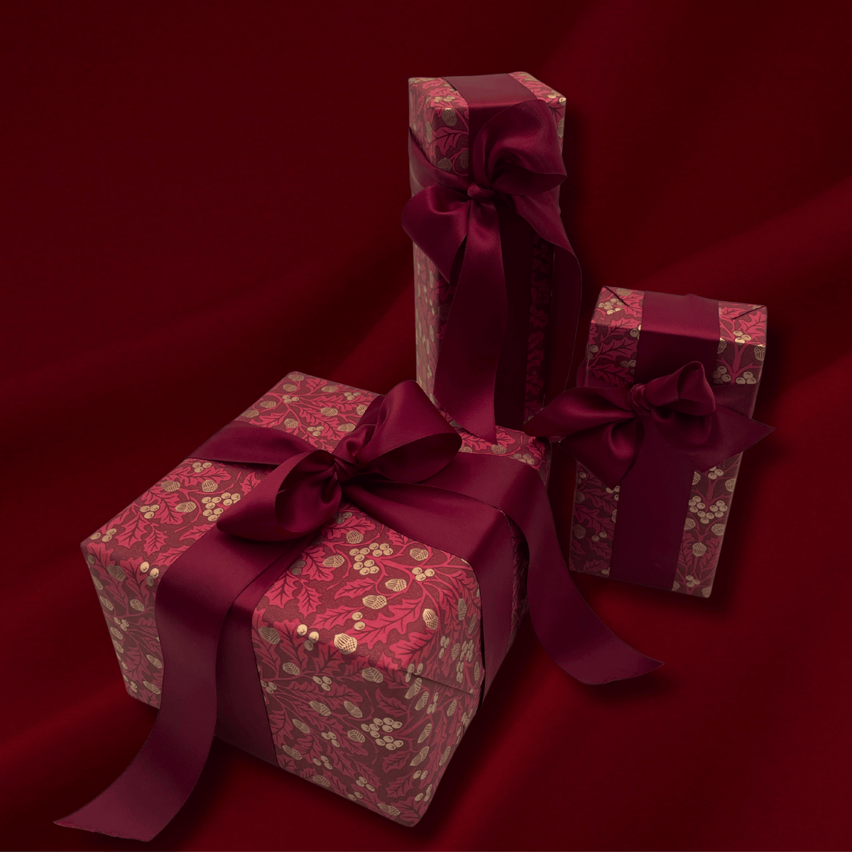 Three wrapped gift boxes with floral patterned wrapping paper and dark burgundy ribbons, arranged on a deep red fabric background.
